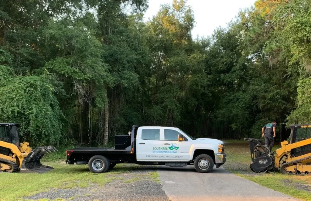 A branded Southern Environmental Premiee Land Management truck and two CAT skid steer loaders, one with a forestry mulching head and operator, staged at a site entrance near dense forest for a major commercial forestry mulching deployment.